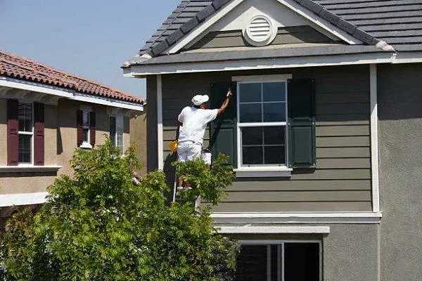 Worker painting the exterior of a gray two-story house on a ladder