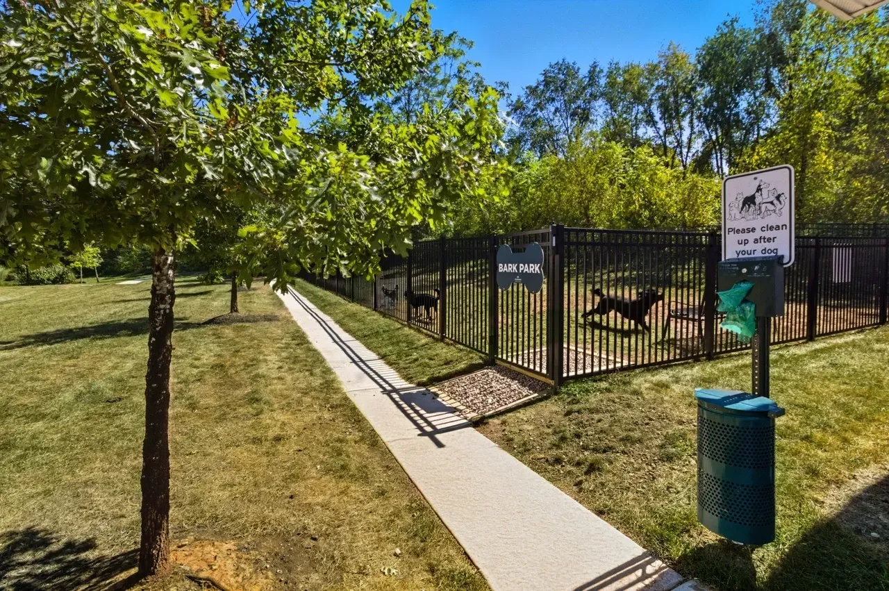 A fenced-in dog park with a paved walkway leading to it, featuring a trash can and dog waste station.