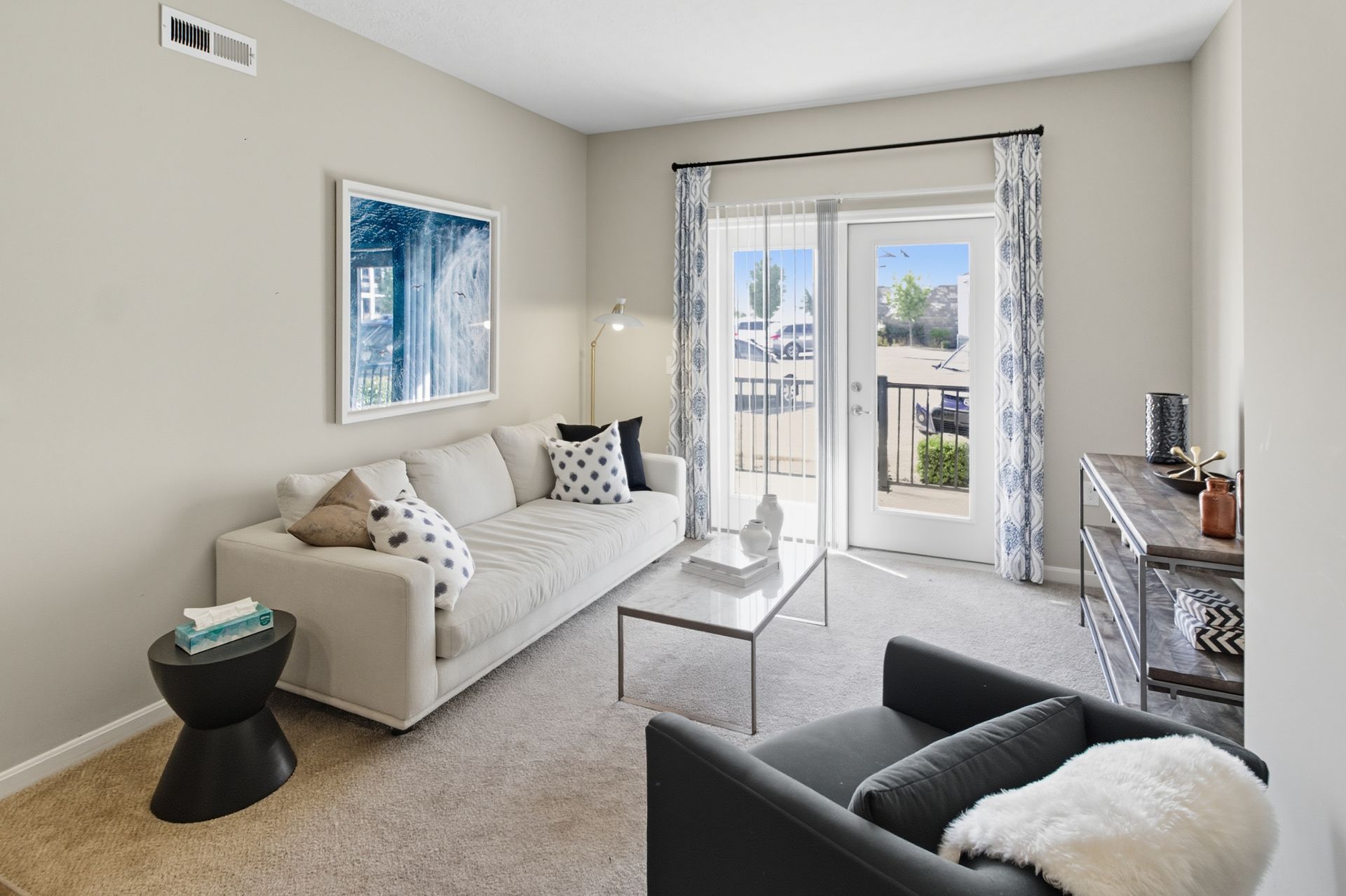 A bright, modern living room featuring a cream sofa, a coffee table, a dark armchair, and a glass door leading outside.