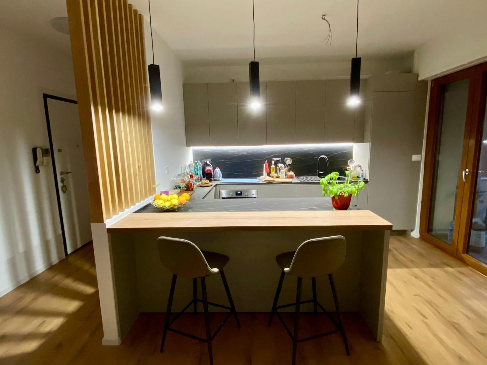 Cucina moderna con isola in legno, due sgabelli, lampade a sospensione e fiori sul bancone.