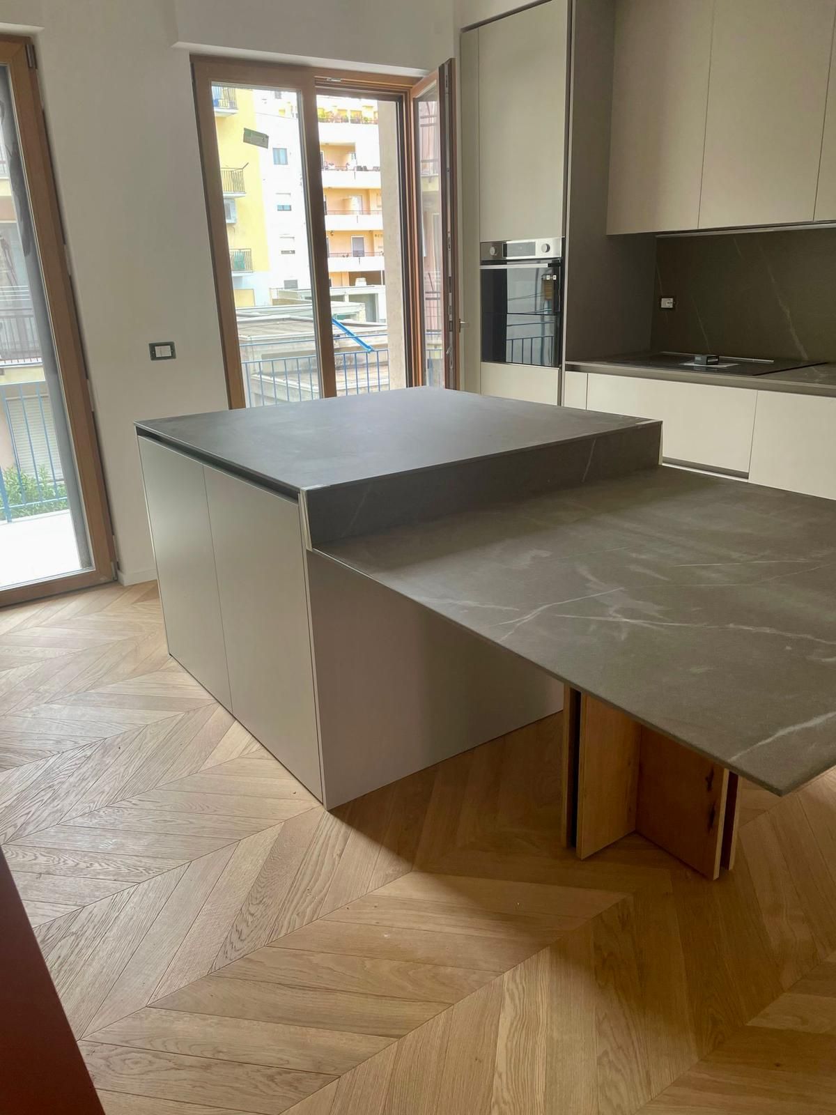 Cucina con isola con piano di lavoro grigio, tavolo estraibile, mobili dai colori chiari e pavimento in parquet.