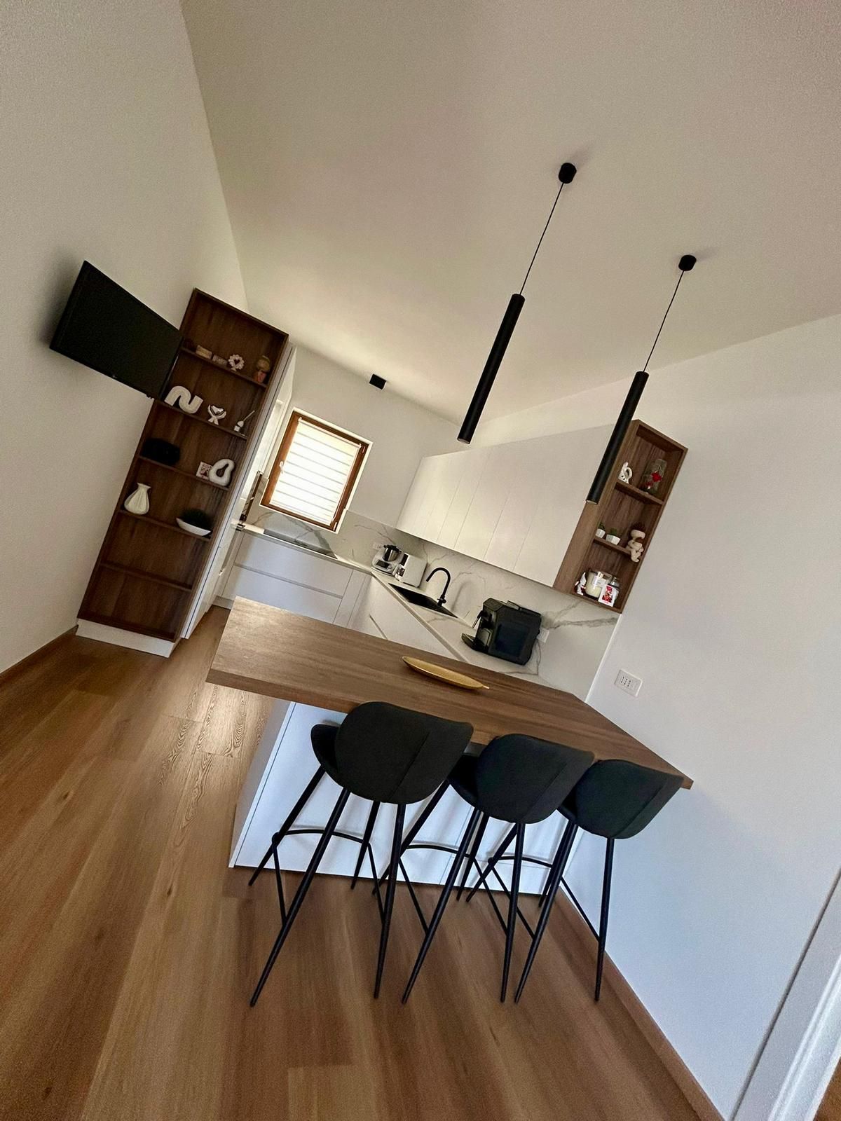 Cucina moderna con isola bianca, sgabelli da bar neri, pavimento in legno e lampade a sospensione.