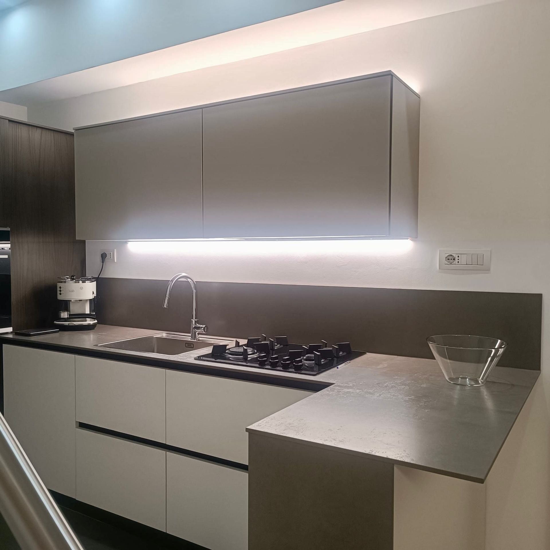 Cucina moderna e minimalista con mobili grigi, piano cottura in acciaio inox e illuminazione sottopensile.