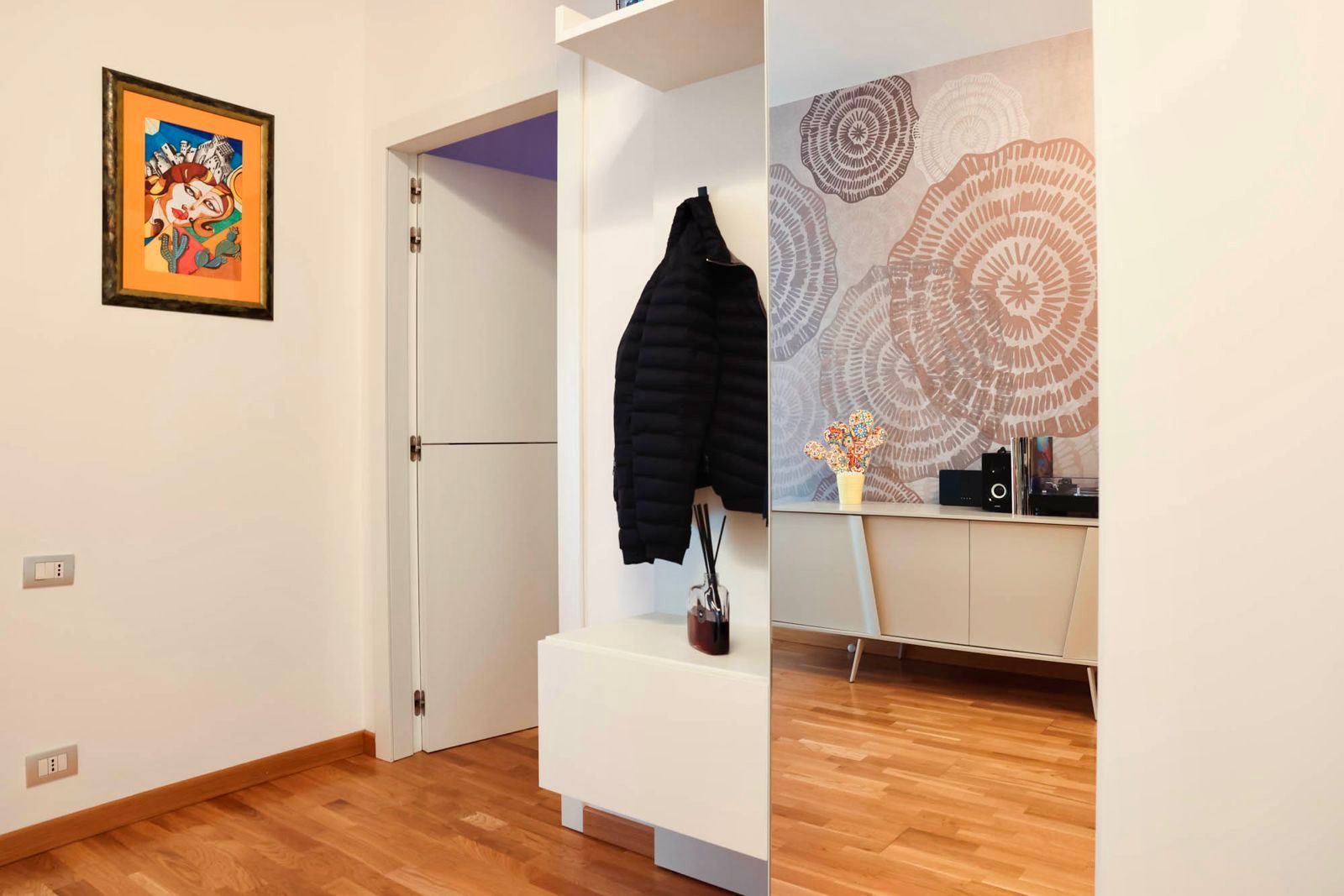 Corridoio moderno con pavimento in legno, appendiabiti, quadri alle pareti, specchio e una porta che conduce a una stanza luminosa.