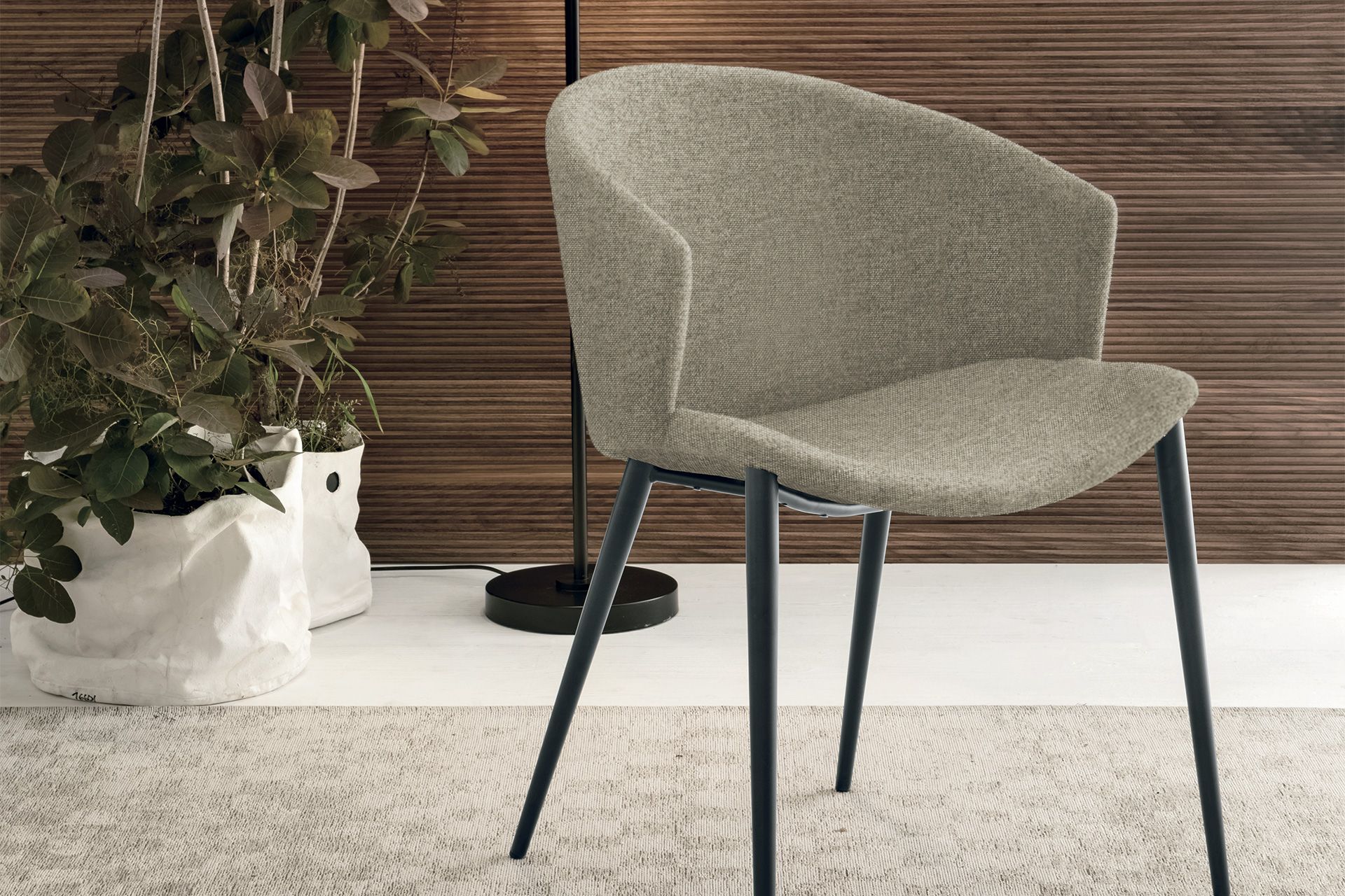 Sedia imbottita grigia con gambe nere, in un ambiente interno moderno con una pianta e una parete rivestita in legno.