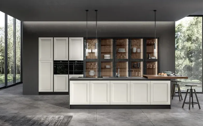 Cucina moderna con mobili bianchi, isola scura, vetrine con ante in vetro e grandi finestre con verde.