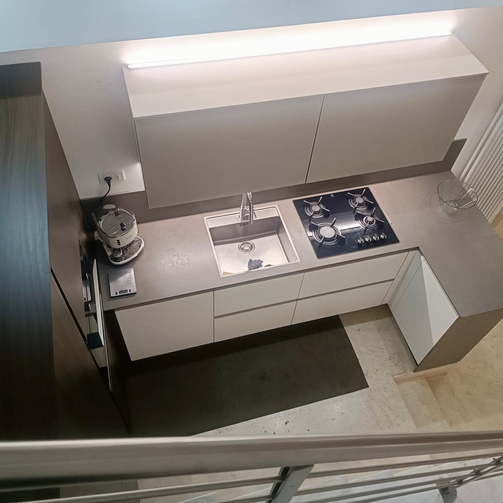 Cucina moderna con mobili bianchi, lavello in acciaio inox e piano cottura a gas sotto una luminosa illuminazione a soffitto.