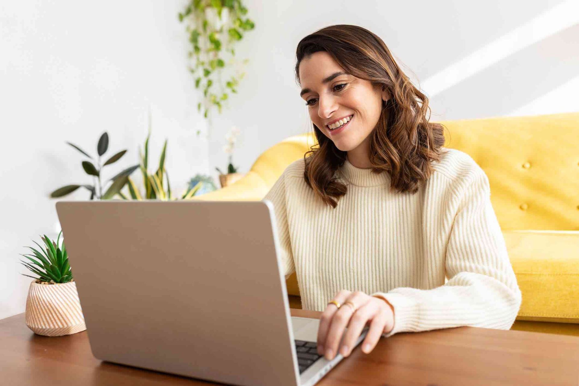 Woman smiling, using a laptop at a table, yellow couch behind her while having a online therapy session.