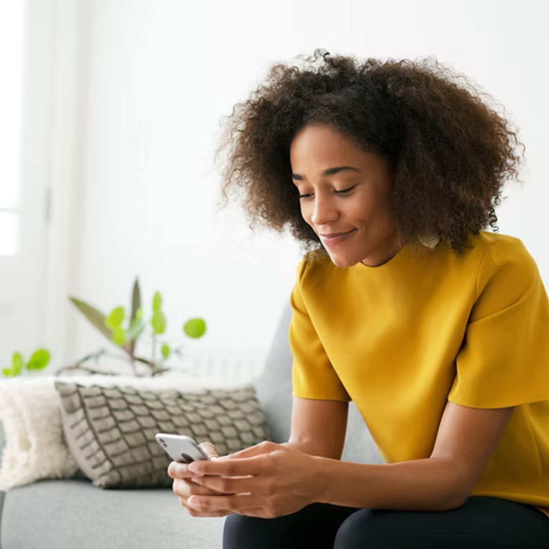 Woman with curly hair in yellow shirt smiles at a phone on a couch indoors while talking with a therapist remotely.