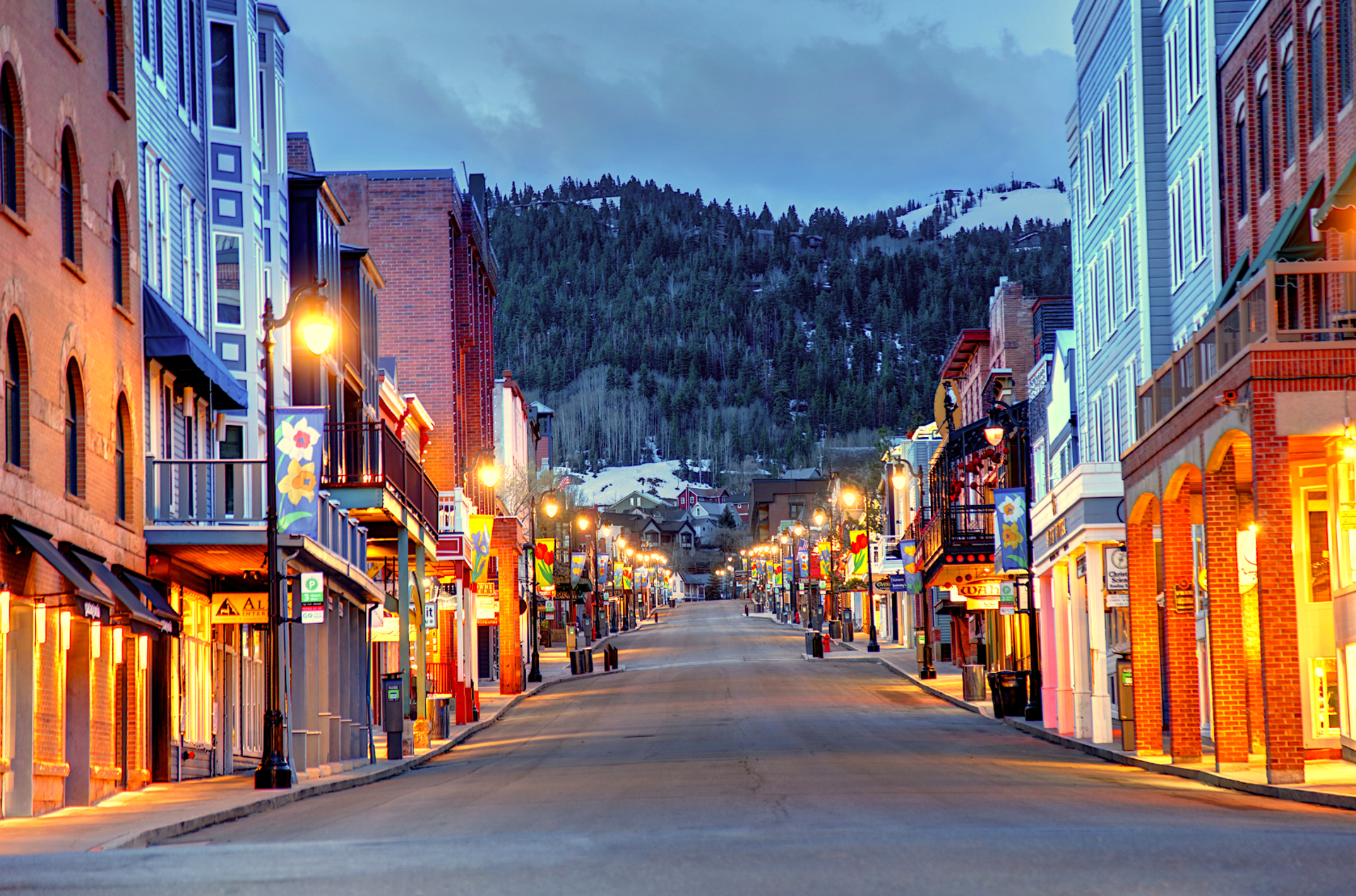 Street lined with buildings and shops, lights on. Mountain in background in Park City. Dusk.