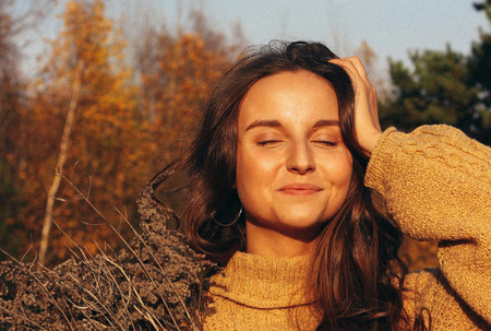 Woman with closed eyes smiles, touching her hair; wearing a yellow sweater, fall foliage background.