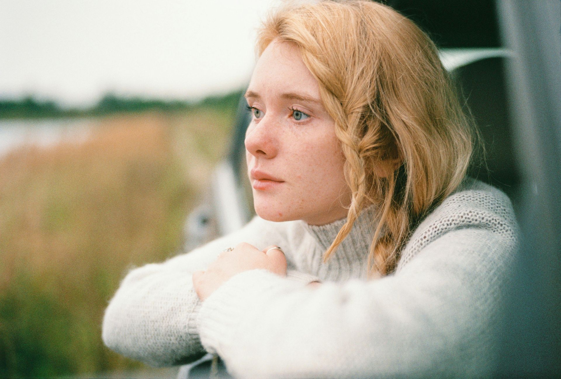 Woman with blonde hair looks out car window, resting arms on the sill, a serene expression. Overcast, outdoor setting.