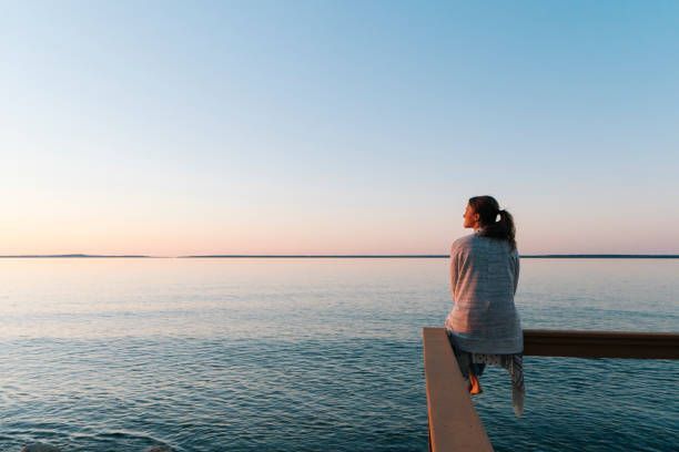 Woman sitting on wooden railing, looking at calm water and sunset. Depression can be heavy. Reset and Rise can help carry that burden with you.