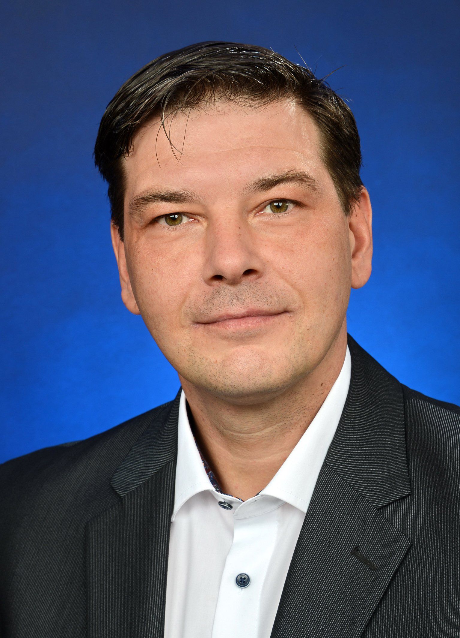 Ein Mann in Anzug und weißem Hemd steht vor einem blauen Hintergrund.