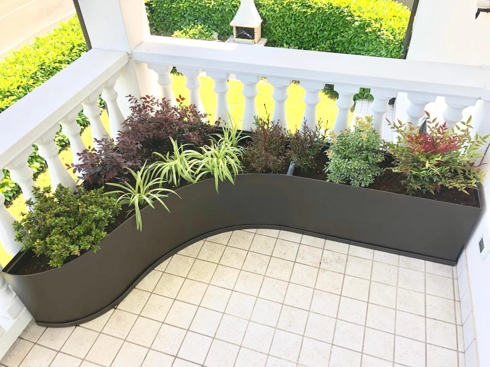 Fioriera nera curva riempita di piante verdi e bordeaux su un balcone piastrellato.