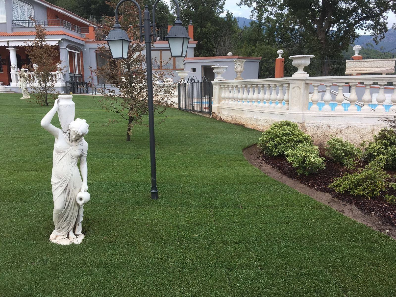 Statua su un prato ben curato accanto a una recinzione bianca e a un giardino, con un edificio sullo sfondo.