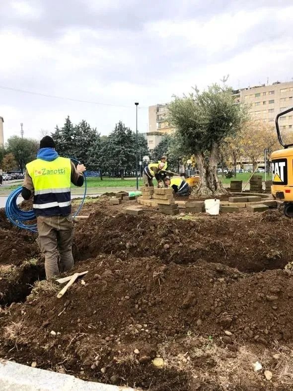 Operai con giubbotti fluorescenti scavano un'aiuola fangosa in un parco, con un piccolo escavatore nelle vicinanze.