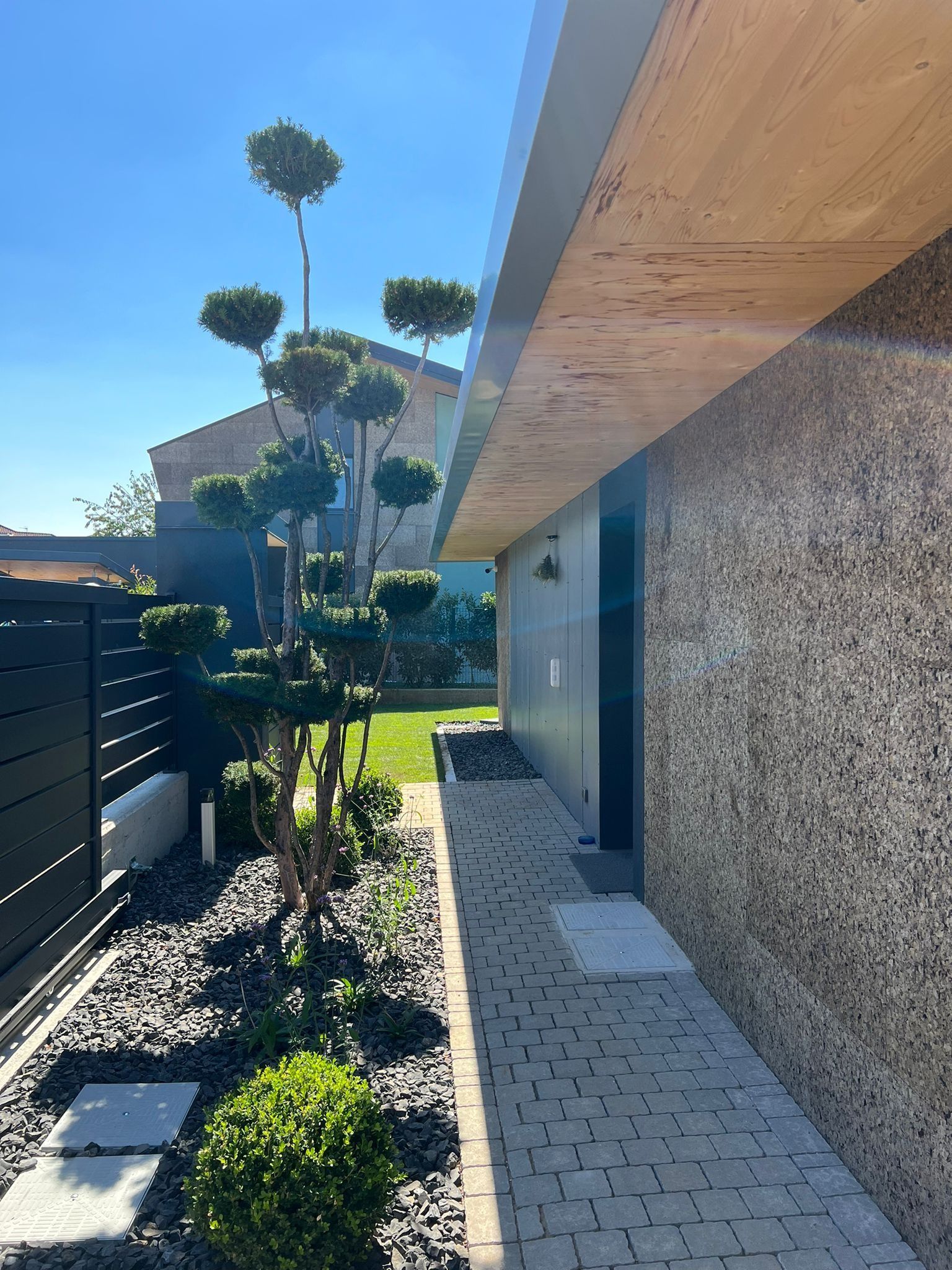 Esterno di una casa moderna con vialetto di ghiaia, arbusti in vaso e cielo azzurro lungo un muro intonacato.