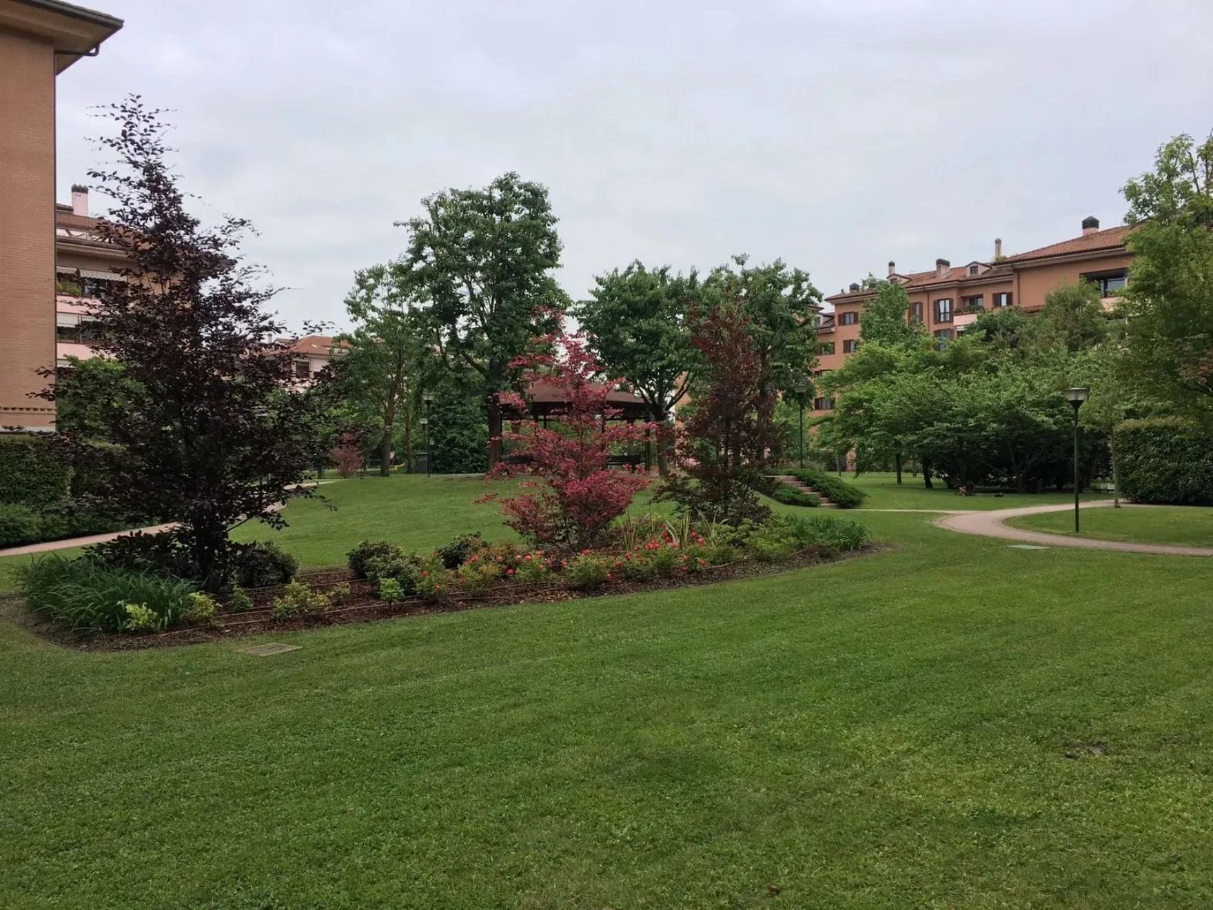 Un prato verde in un parco con alberi, arbusti e palazzi residenziali sullo sfondo.