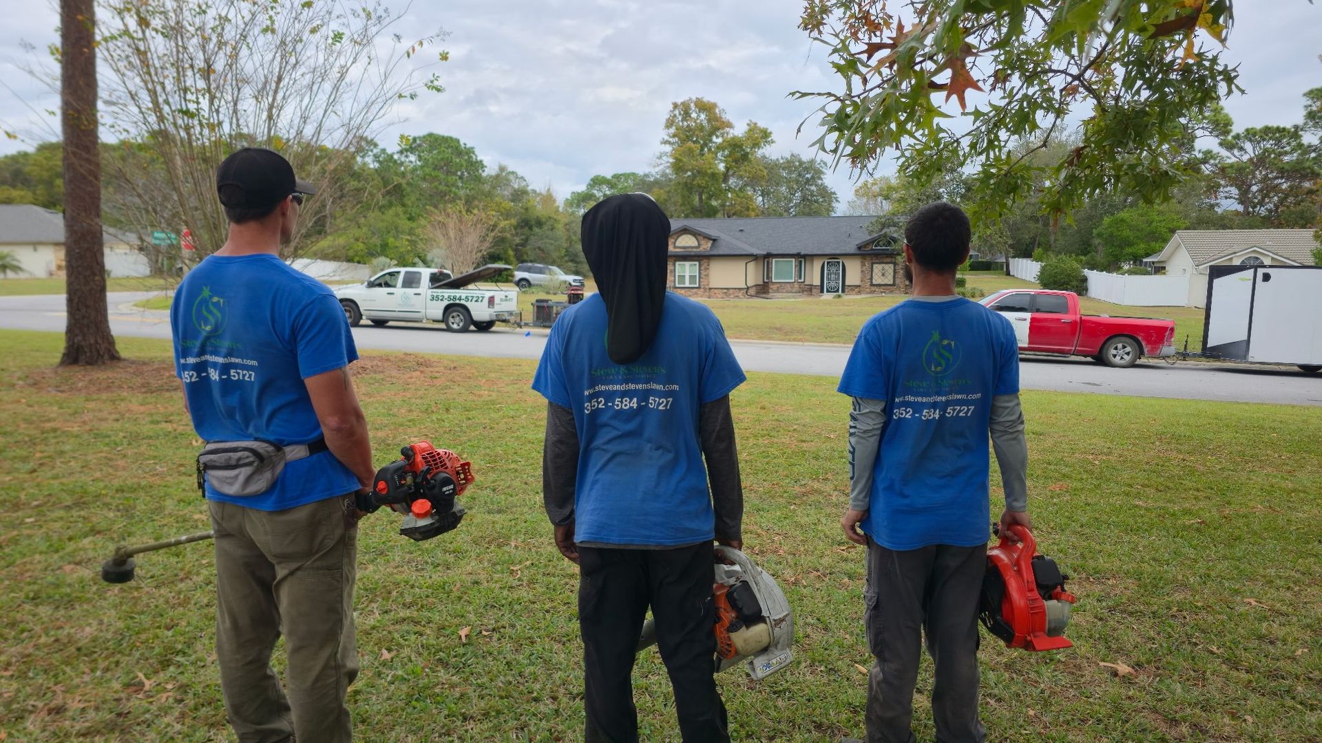Three people in blue shirts with yard tools, standing on grass in front of a house.