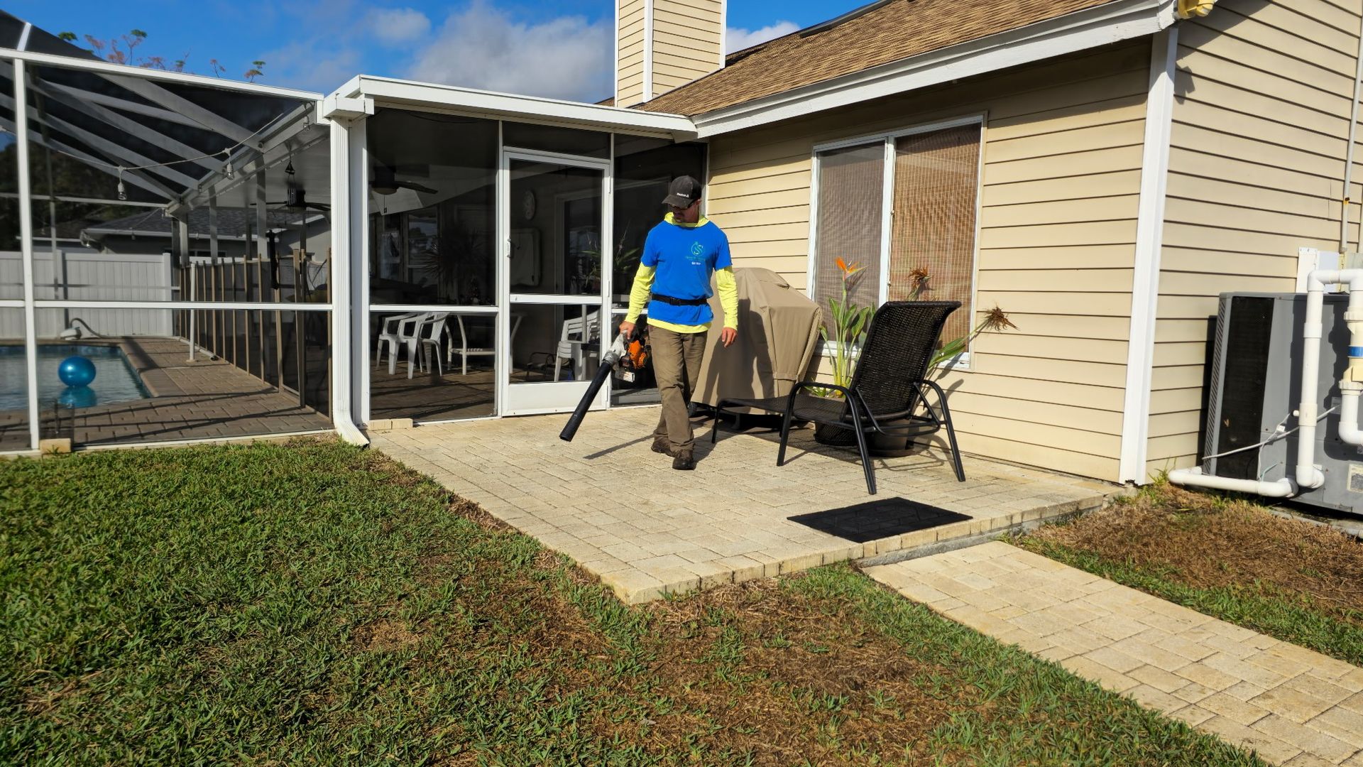 Person using a leaf blower on a patio next to a house with a screened pool area.