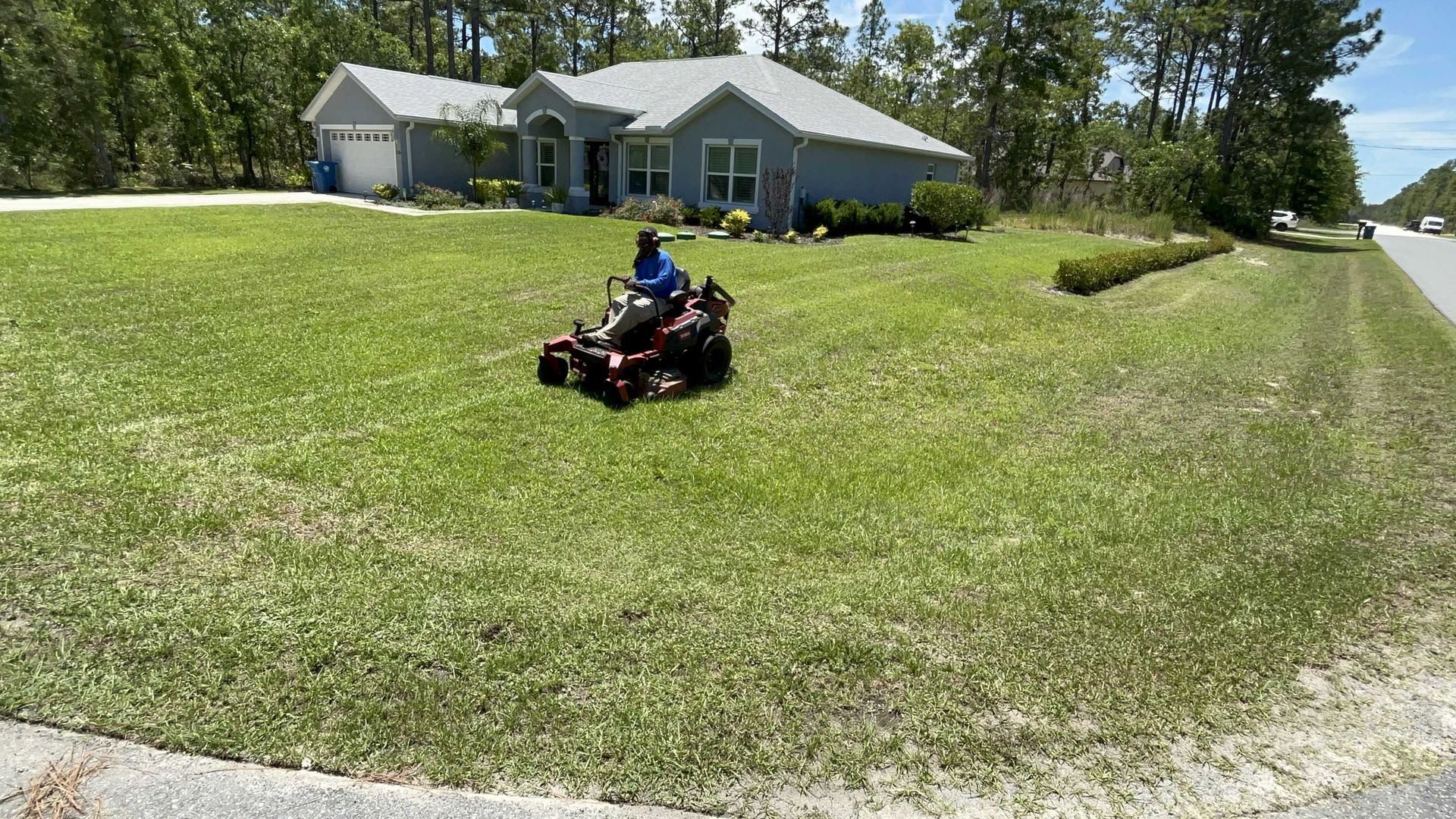 Person mowing a large lawn on a riding mower in front of a blue house on a sunny day.