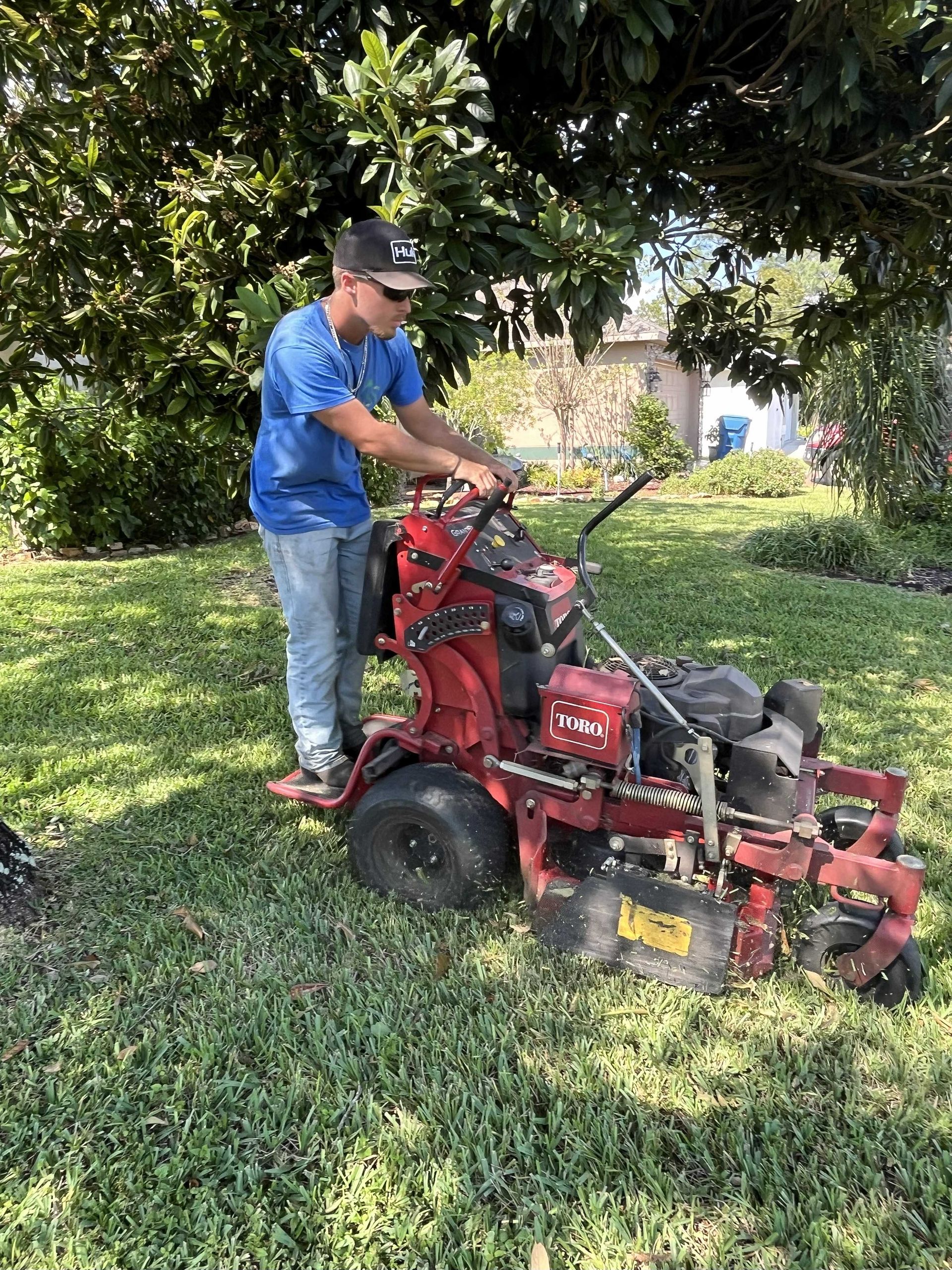 Man operating a red lawn mower on a grassy yard under a tree.