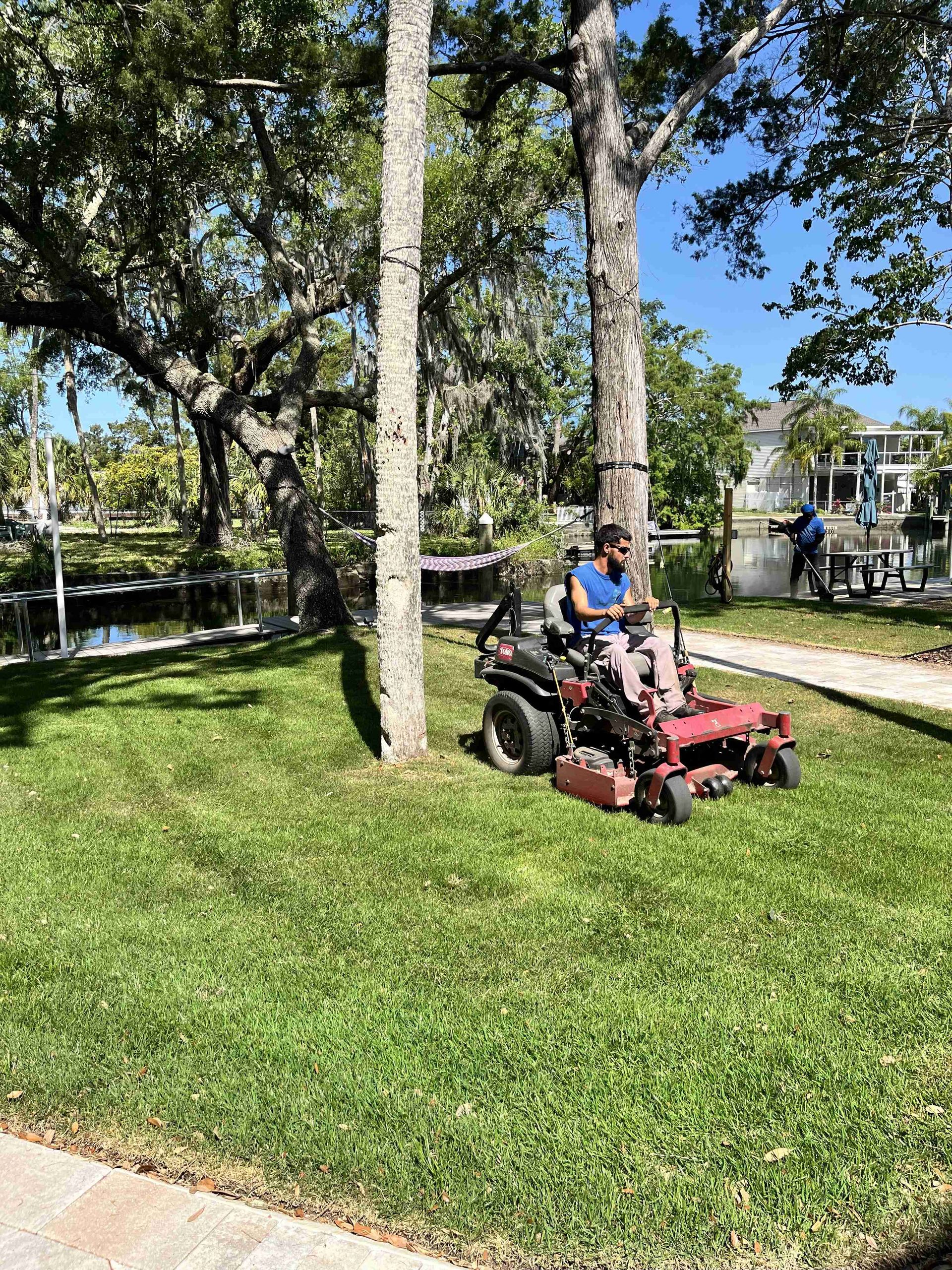 Man on riding lawnmower cutting grass near trees and a canal on a sunny day.