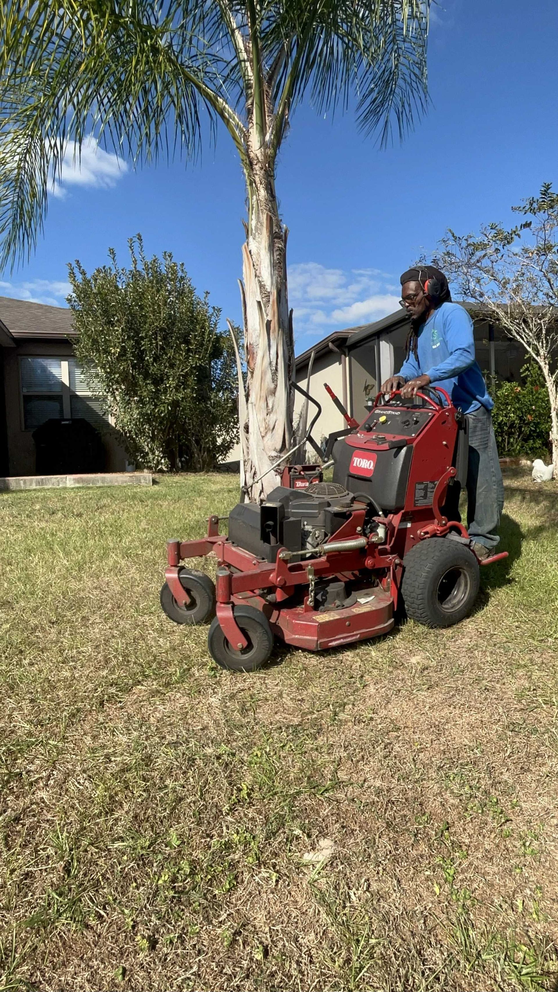 Person operating a red Toro zero-turn mower on a grassy lawn with a tree, sunny day.