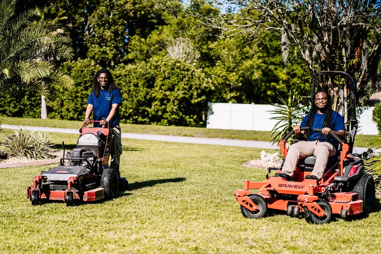 Two people are riding lawn mowers on a lush green lawn.