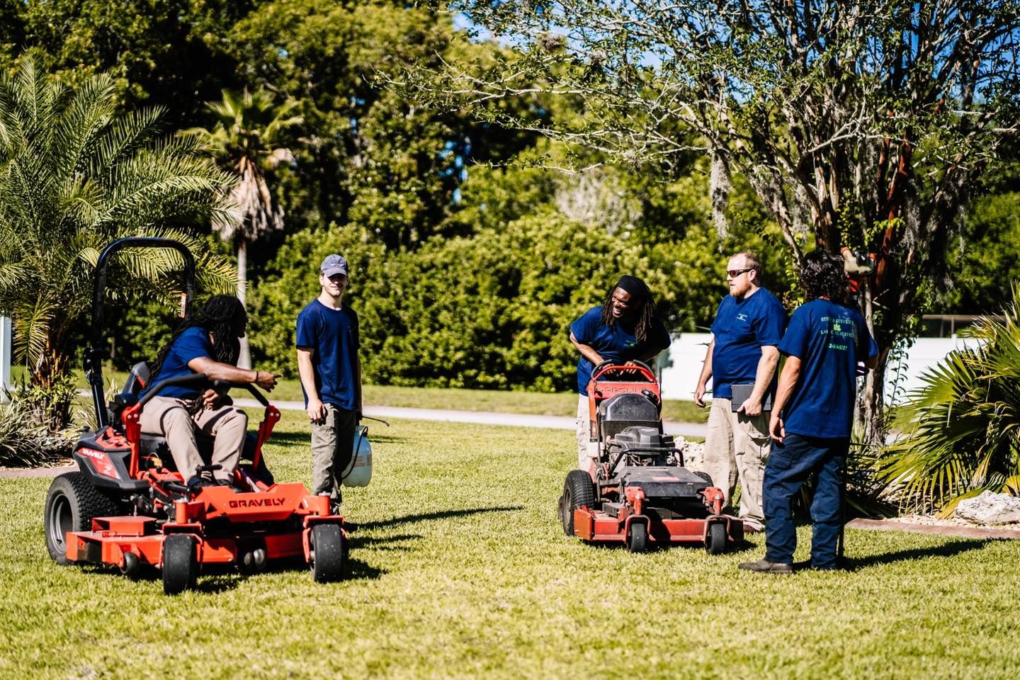 A group of people are standing around lawn mowers on a lush green lawn.