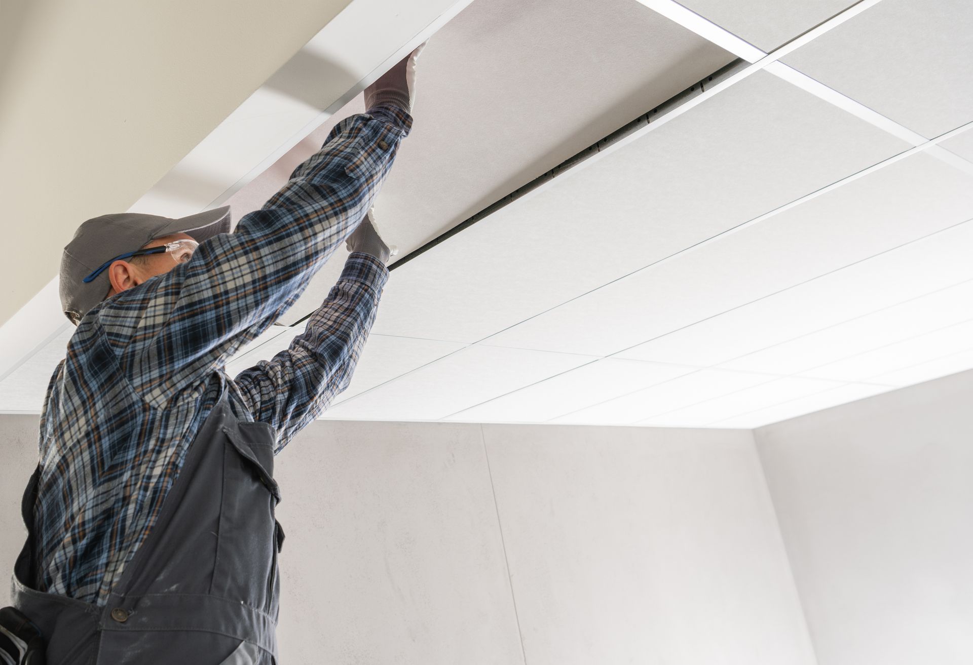 Man in work clothes installing ceiling tiles. Indoors, arms raised, working on a grid ceiling.