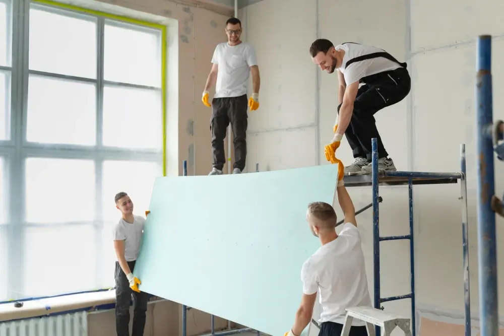 Four construction workers installing drywall in a room with a large window.