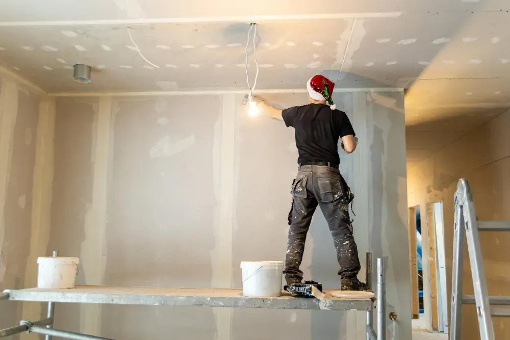 A person wearing a Santa hat works on a wall, standing on scaffolding, wires and buckets visible.