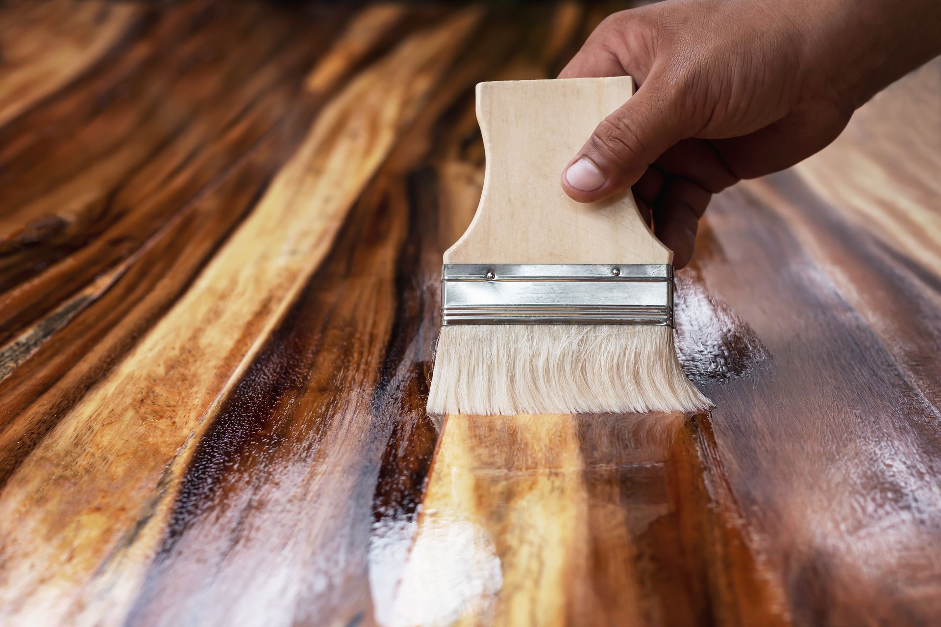 Hand applying varnish to a wood surface with a brush, creating a glossy, wet finish.
