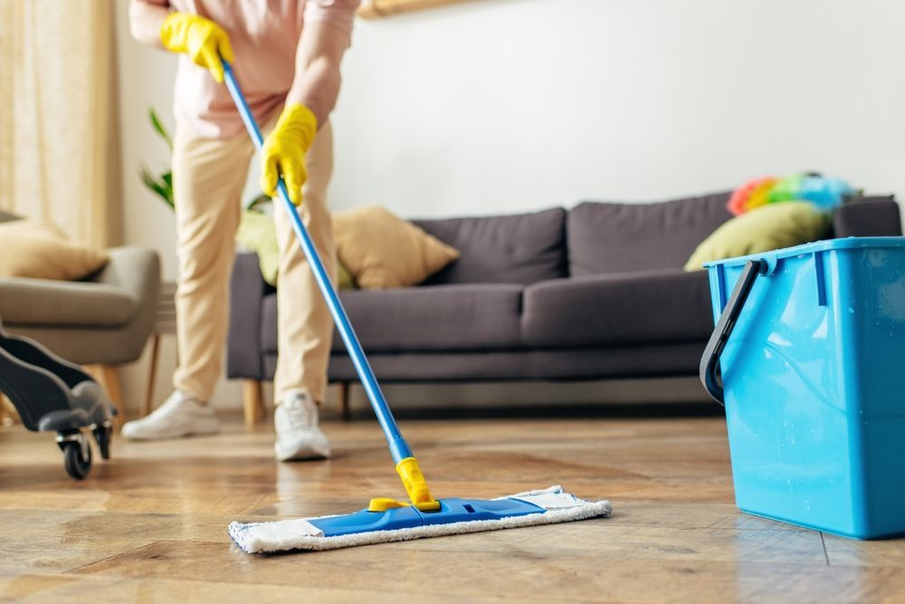 Person mopping a wood floor in a living room, wearing yellow gloves, with a bucket and couch in the background.