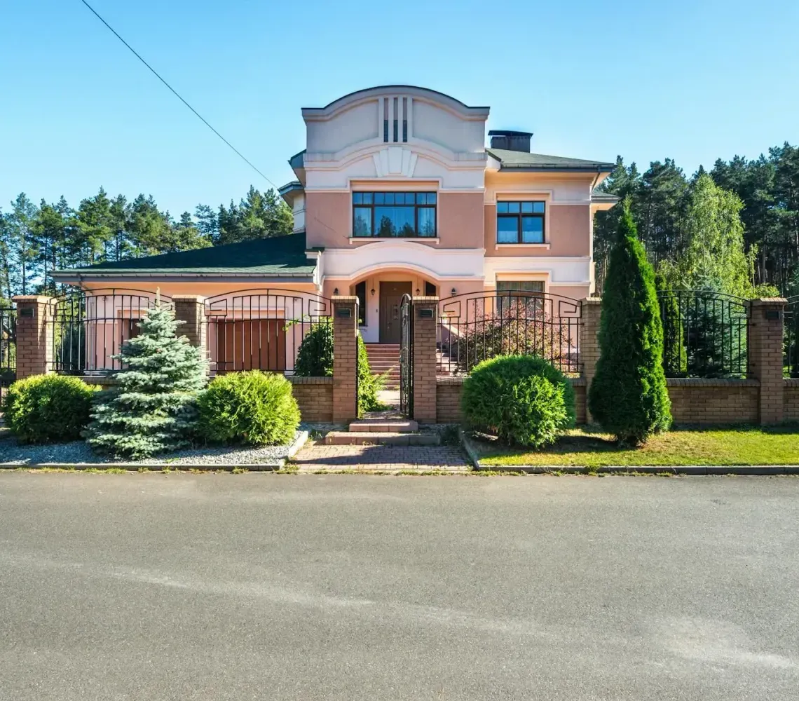 Large peach-colored house with a front gate, landscaped yard, and trees along a quiet street