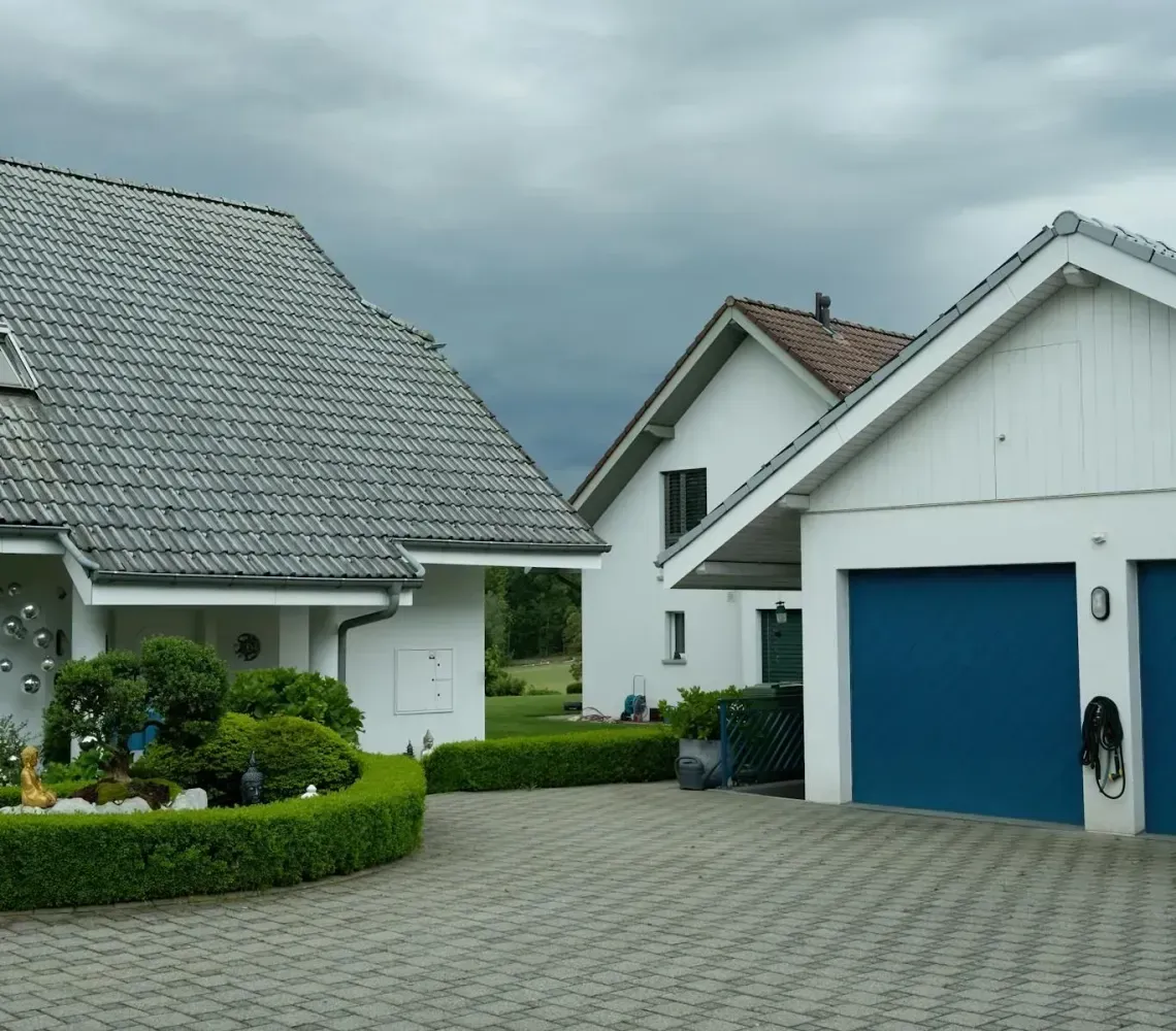 Quiet residential driveway with white houses, gray roofs, blue garage doors, and a cloudy sky