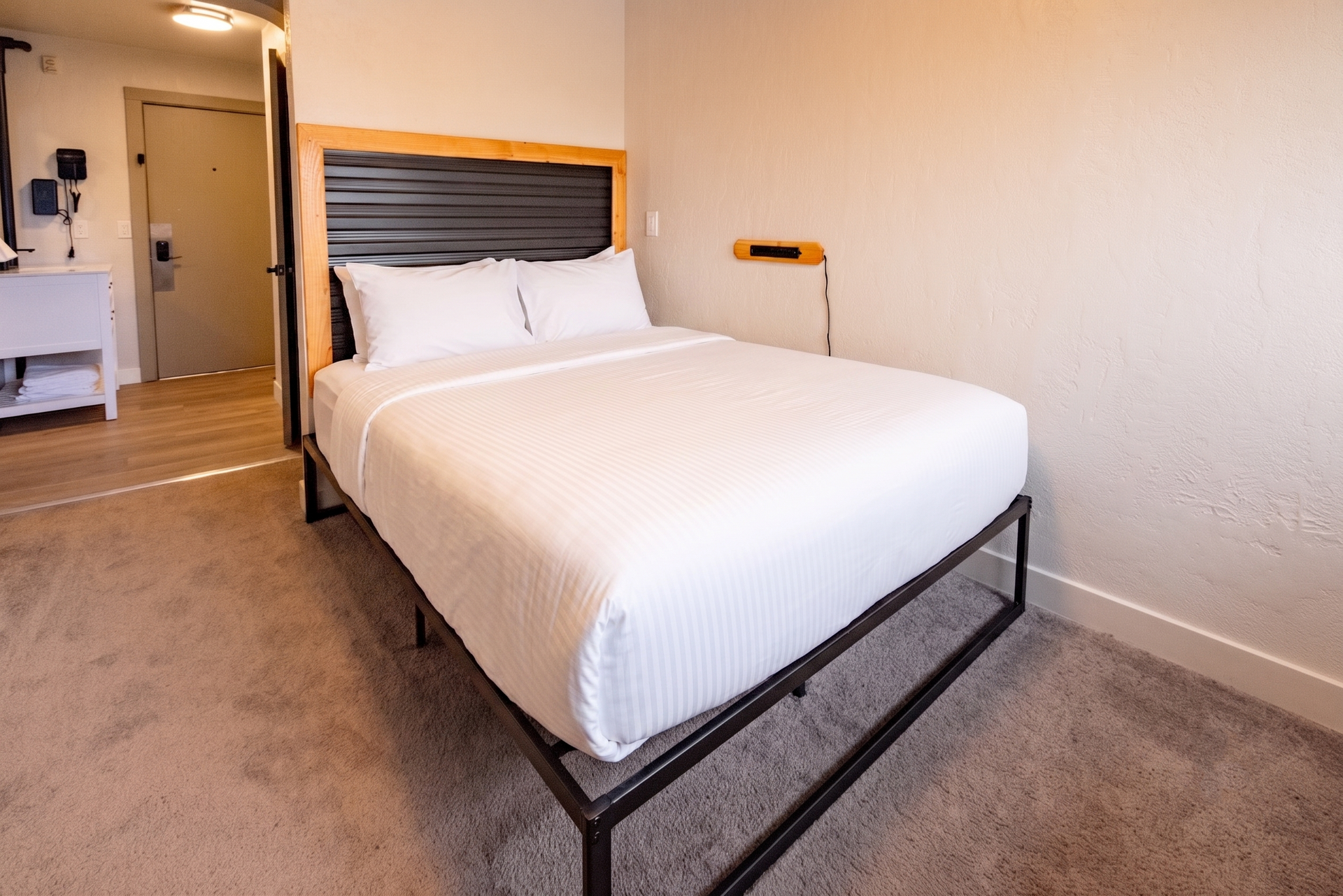 Hotel room with a neatly made bed, beige carpet, and hallway visible in the background