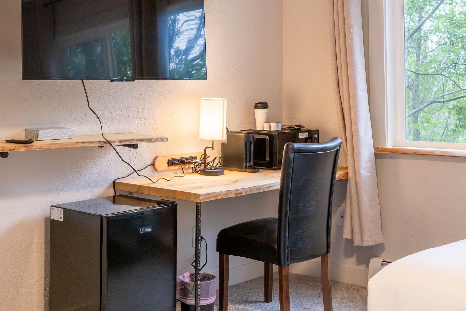 Small desk nook with lamp, chair, mini fridge, coffee maker, and wall-mounted TV by a window.