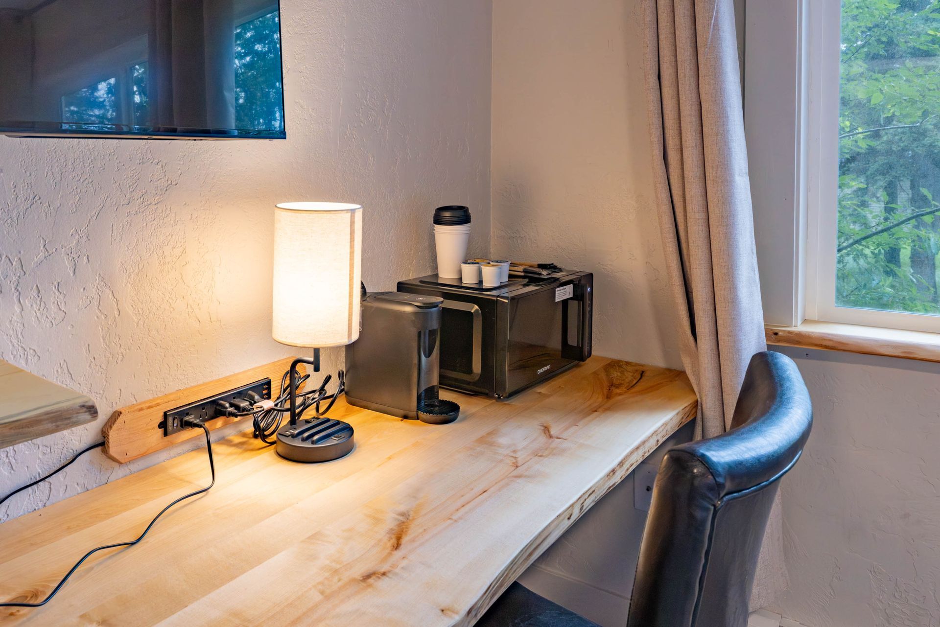 Small desk by a window with a lit lamp, coffee maker, and chair in a home office.
