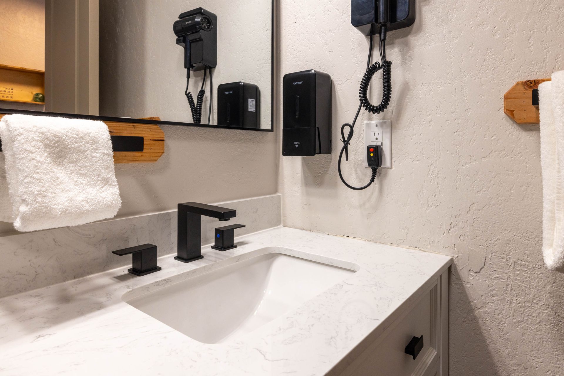 Modern white bathroom sink with black faucet, mirror, towel, soap dispenser, and wall-mounted accessories.