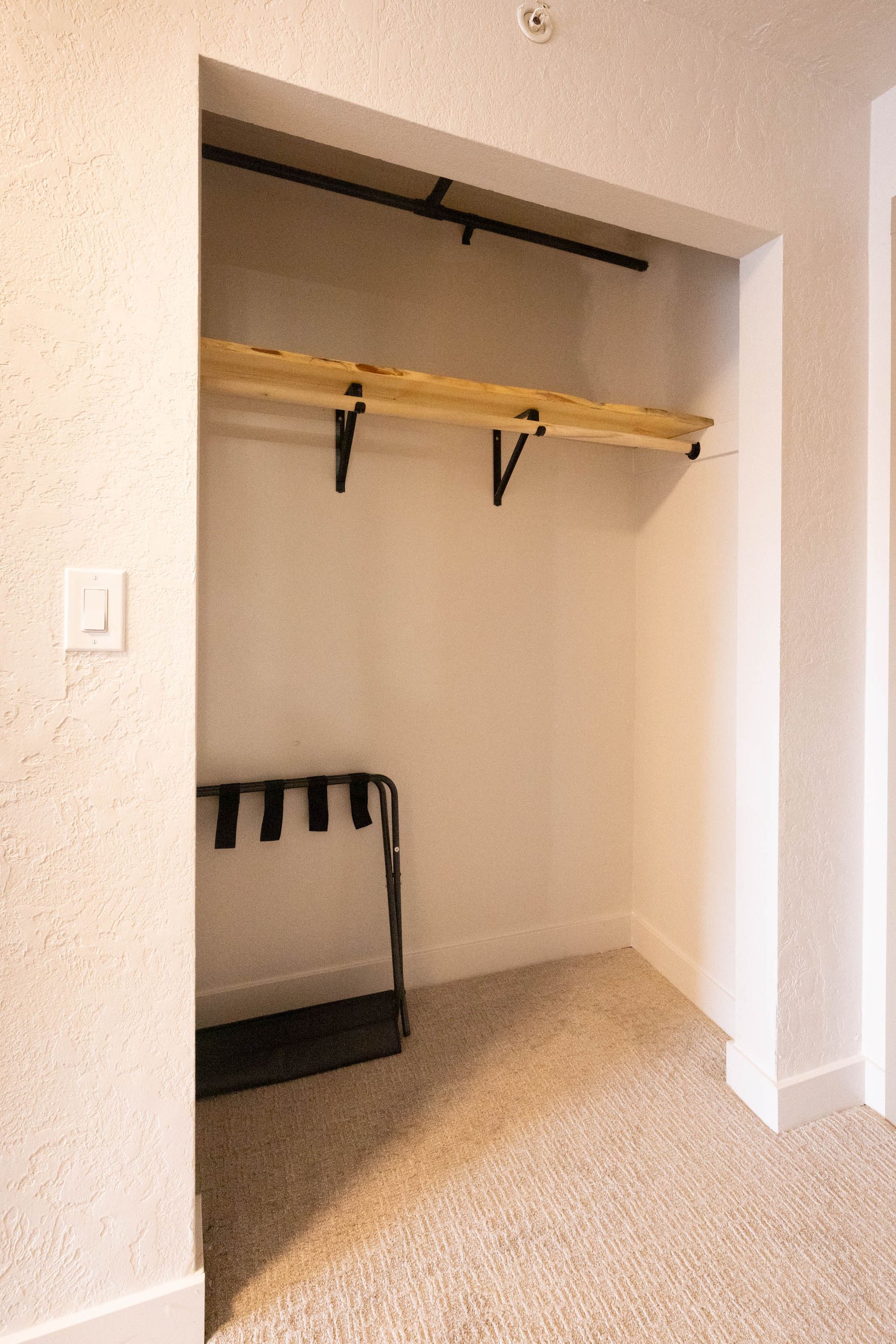 Empty closet with a wooden shelf and black metal rack inside, beige carpet and walls.