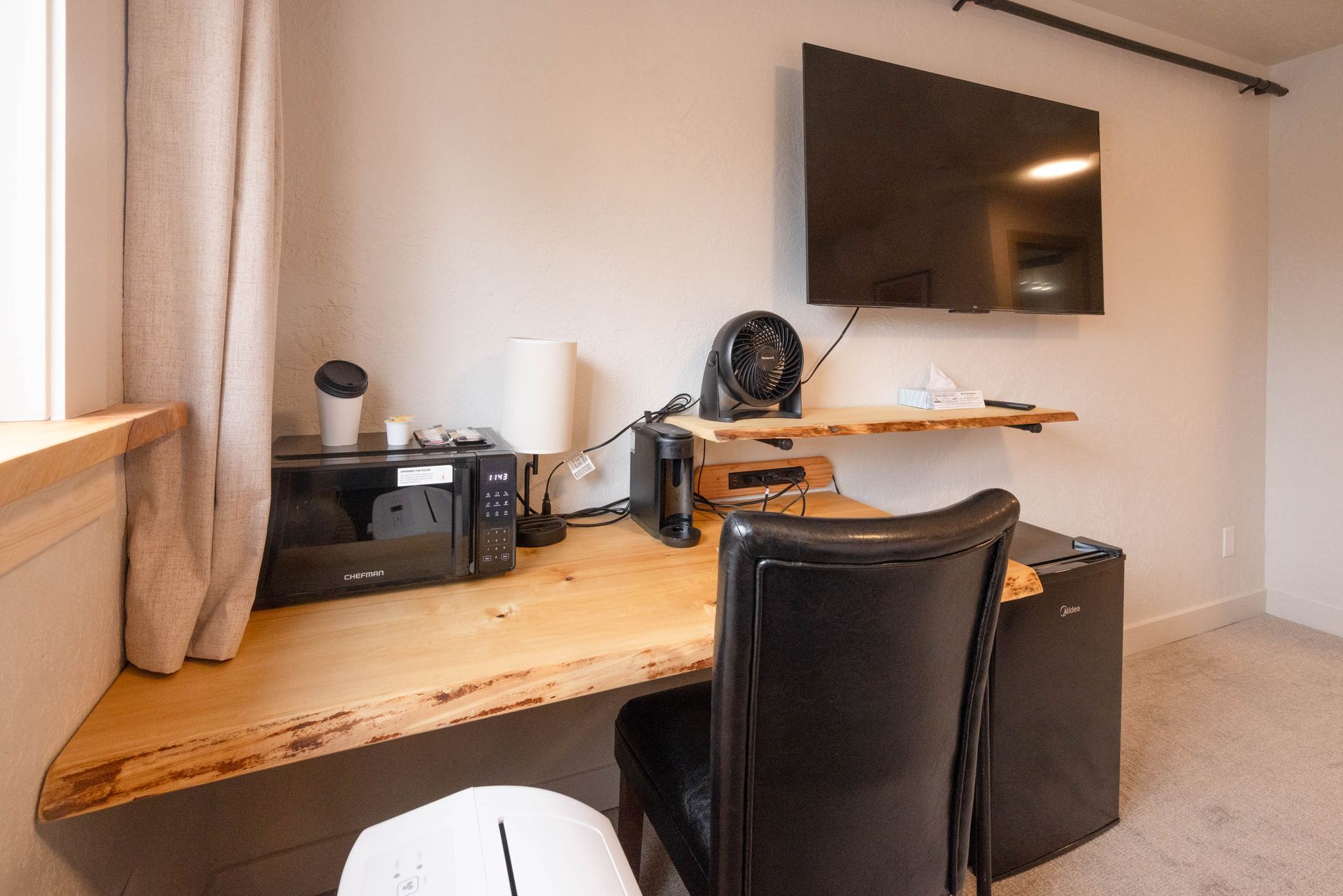 Home office desk with wall-mounted TV, black chair, and beige curtains in a bright room