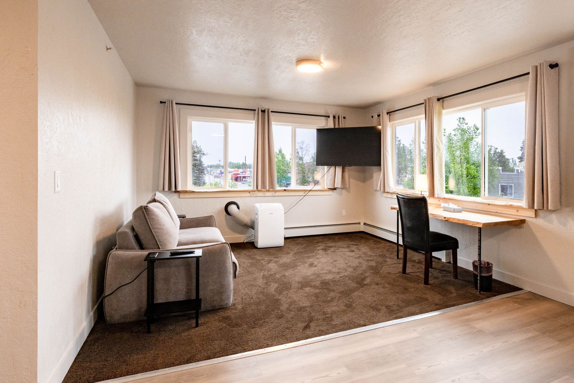 Bright living room with sofa, chair, desk, wall-mounted TV, and large windows.