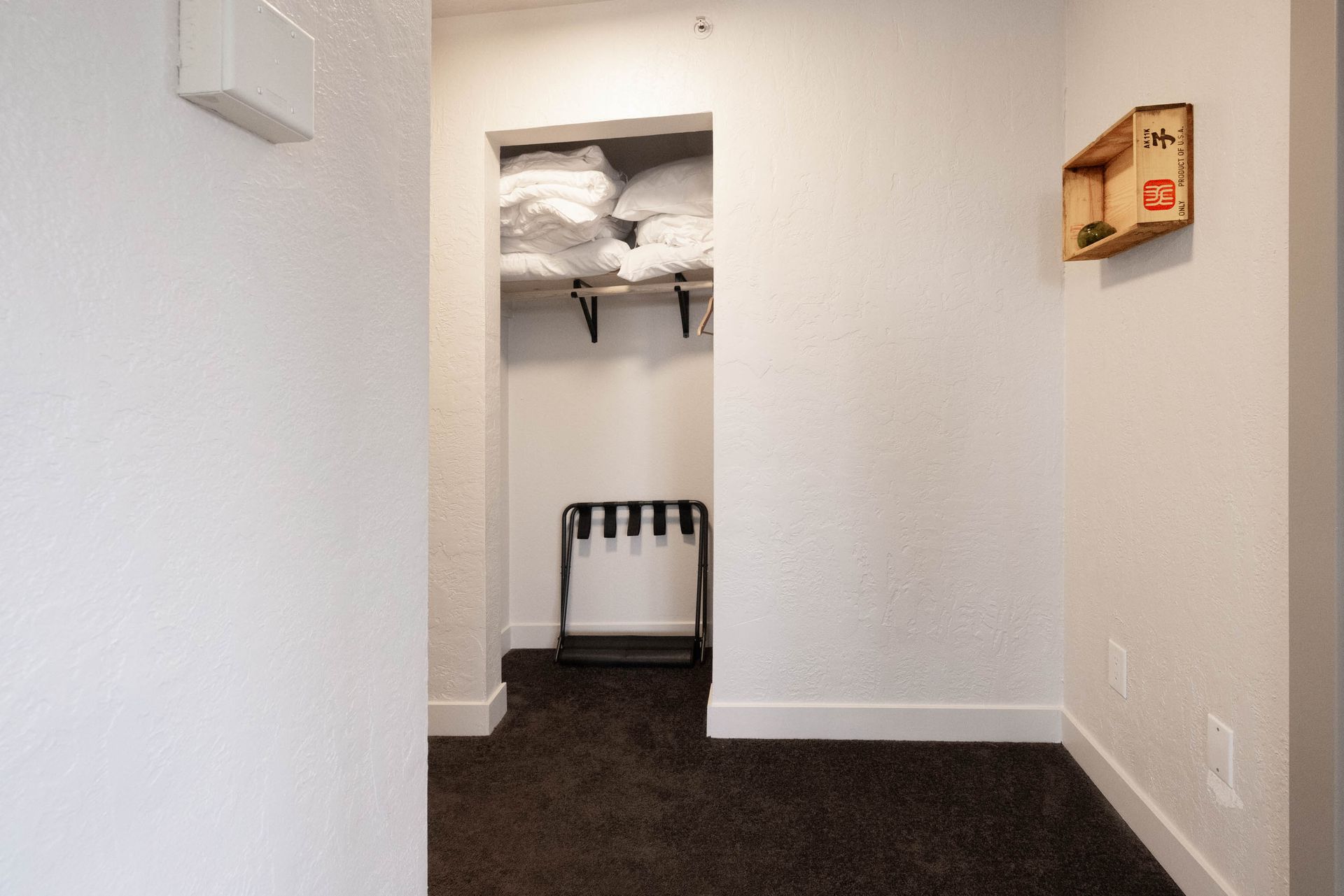 Small hallway with an open closet, folded linens on a shelf, and a wall-mounted box on the right.