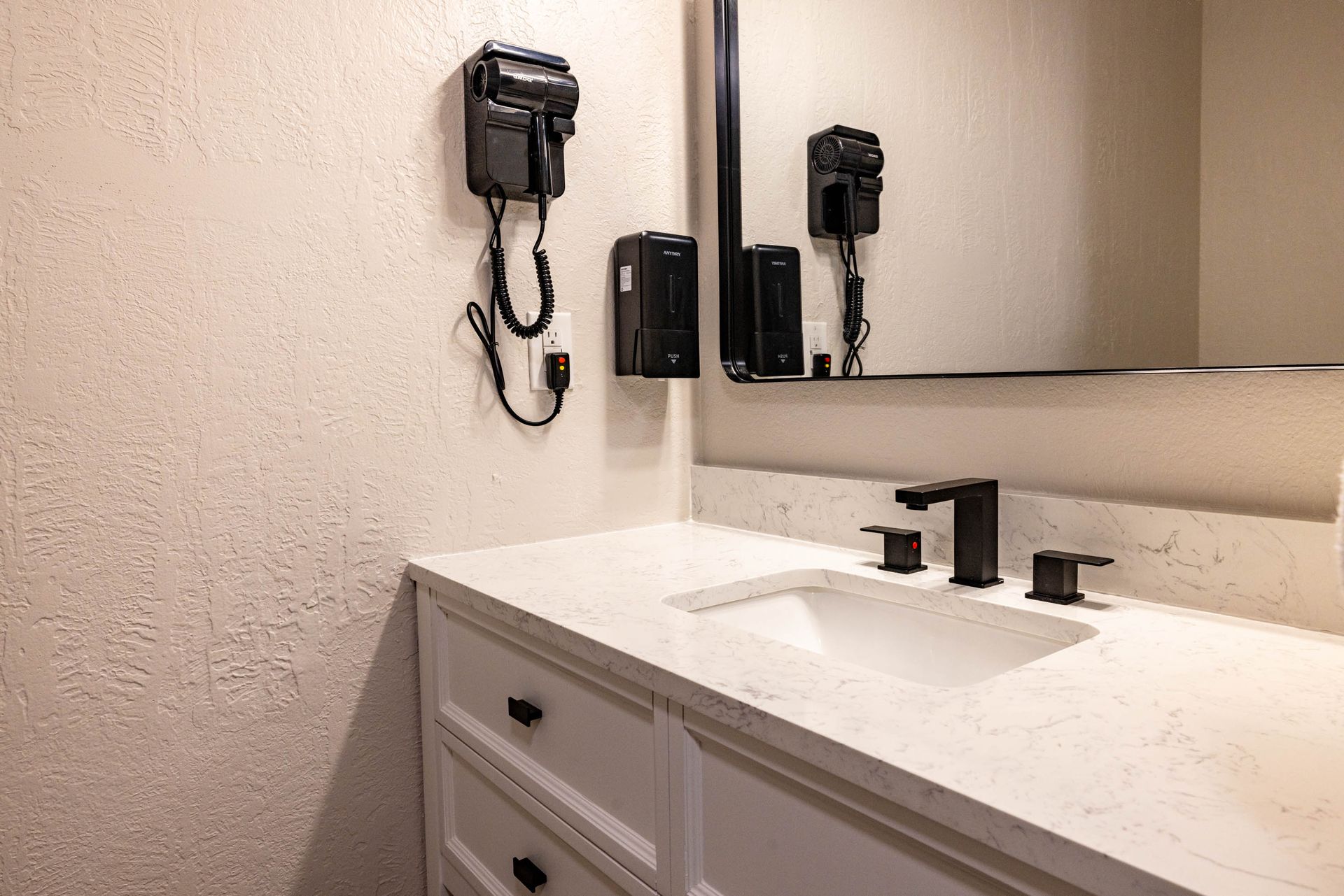 Bathroom vanity with a mirror, sink, soap dispenser, and wall-mounted hair dryer.