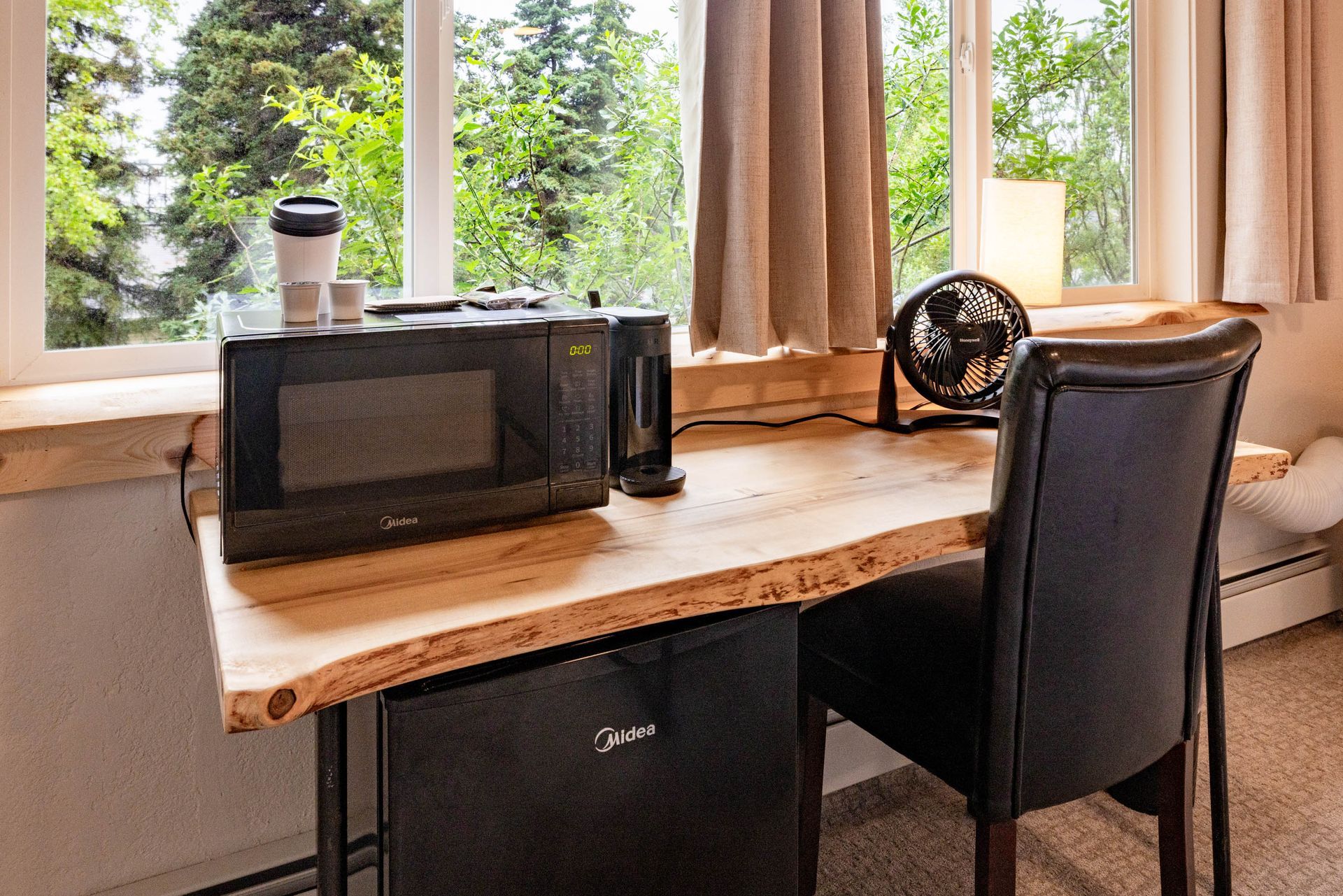 Small desk by a window with a microwave, mini fridge, chair, and lamp in a cozy room