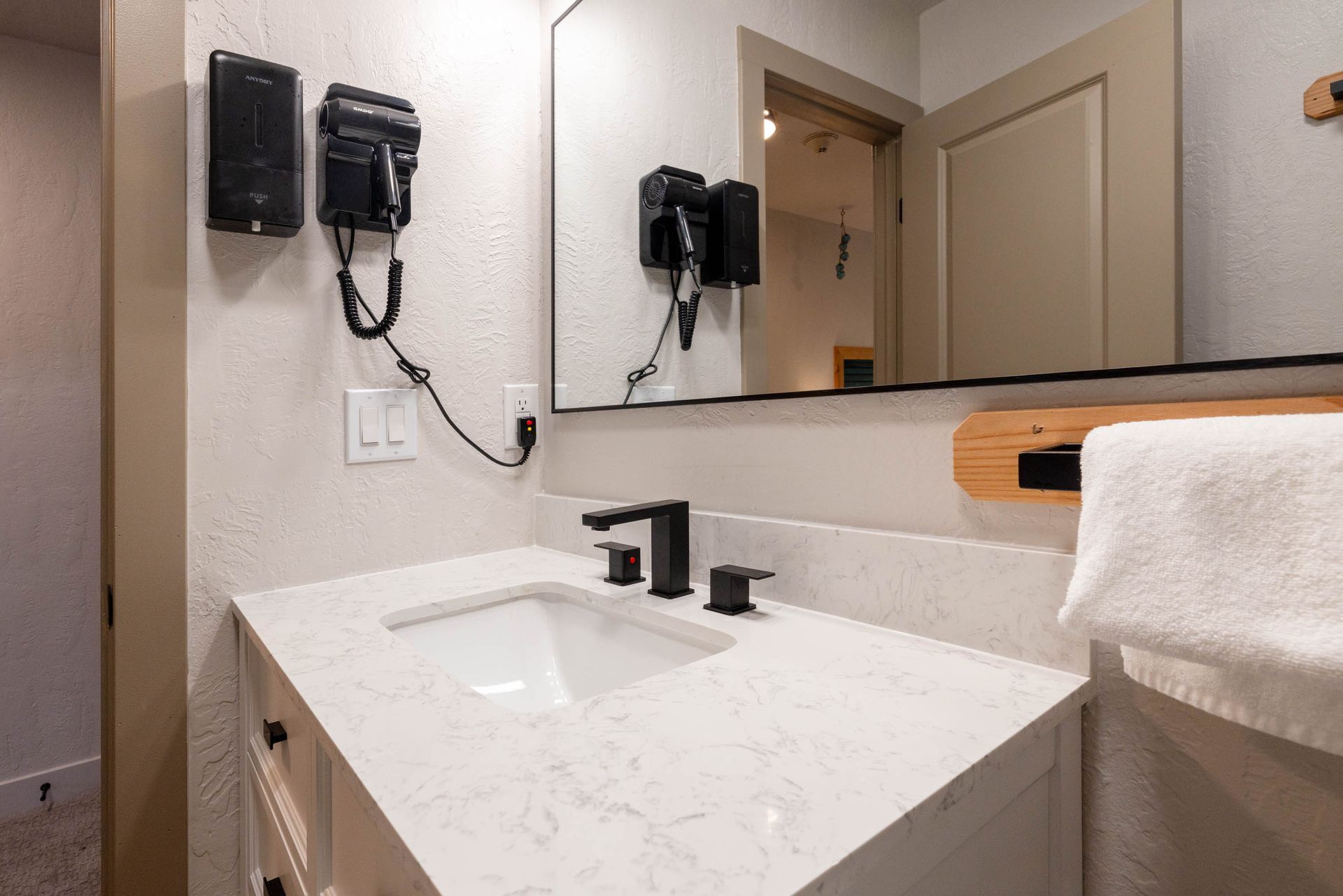 Modern bathroom vanity with white countertop, black faucet, wall-mounted hair dryers, and mirror