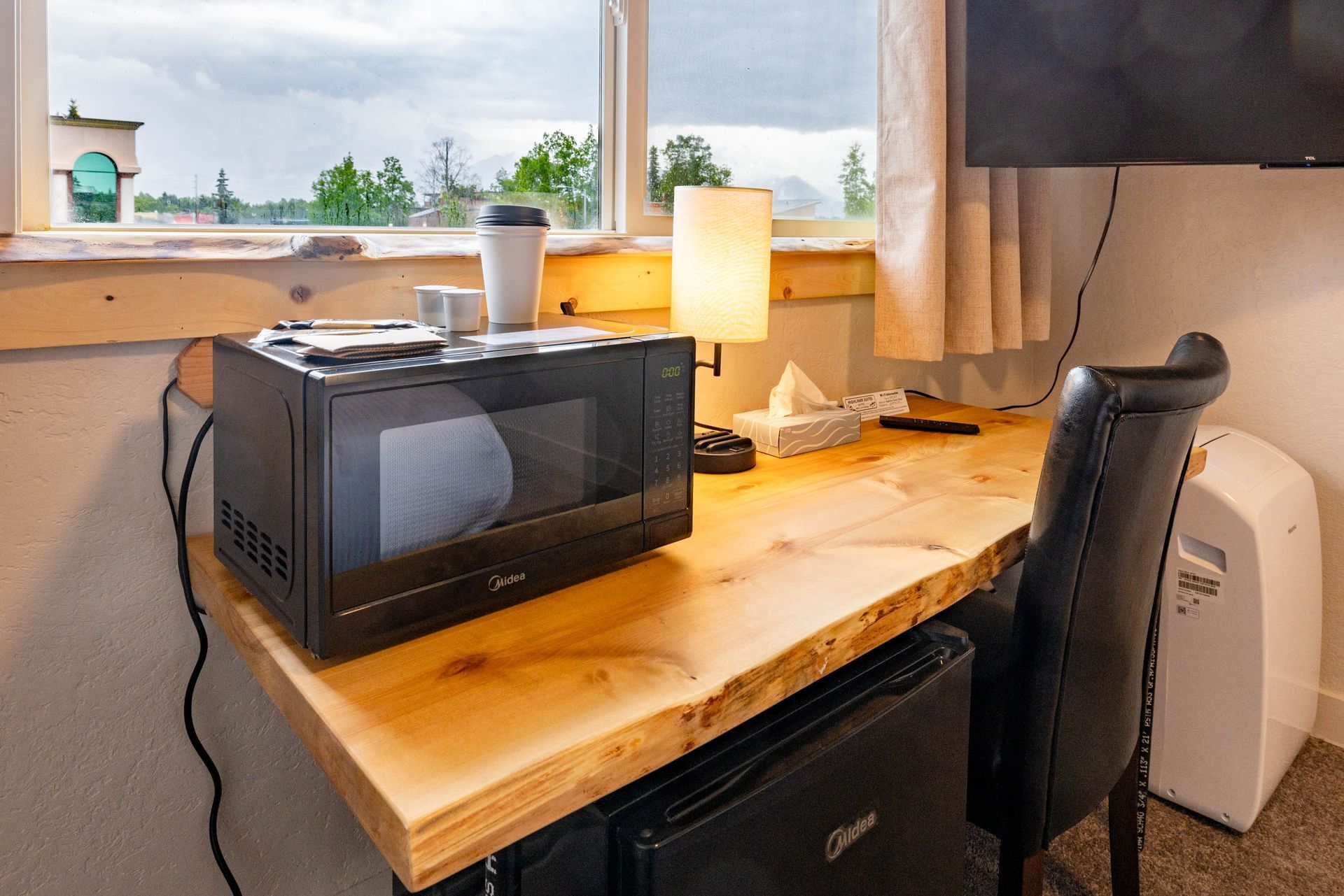 Small office desk by a window with a microwave, lamp, chair, and laptop on a wooden surface.